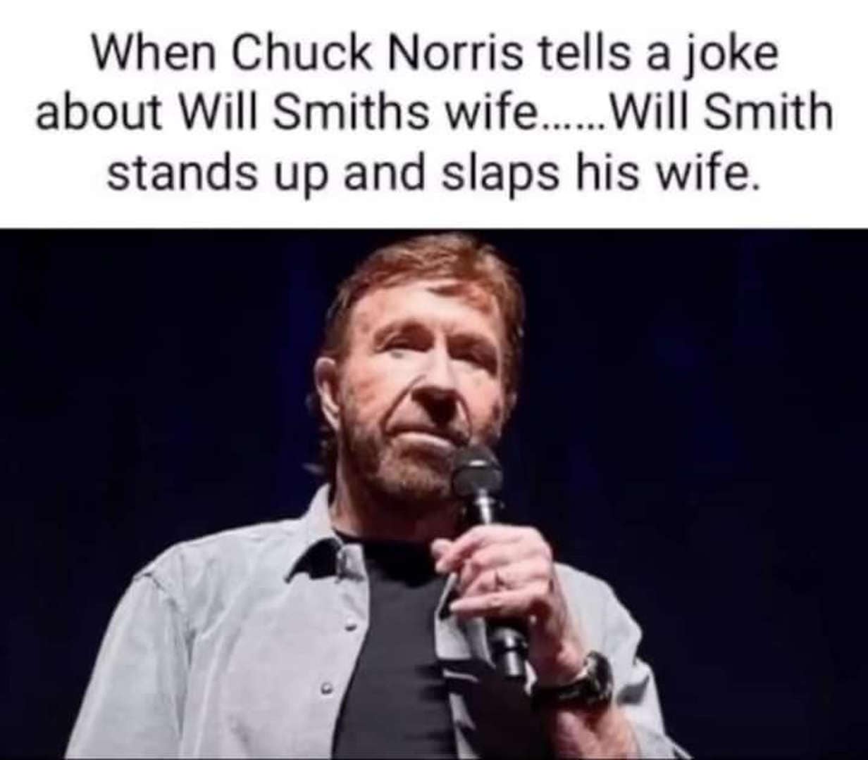 Chuck Norris stands under a spotlight holding a microphone, looking serious. This entry into the world of Chuck Norris memes references pop culture, stating that if Chuck joked about Will Smith's wife, Will would simply slap his own wife rather than confront the legend.