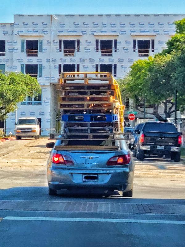 A driving fail showing a silver sedan carrying a dangerously high stack of wooden pallets.