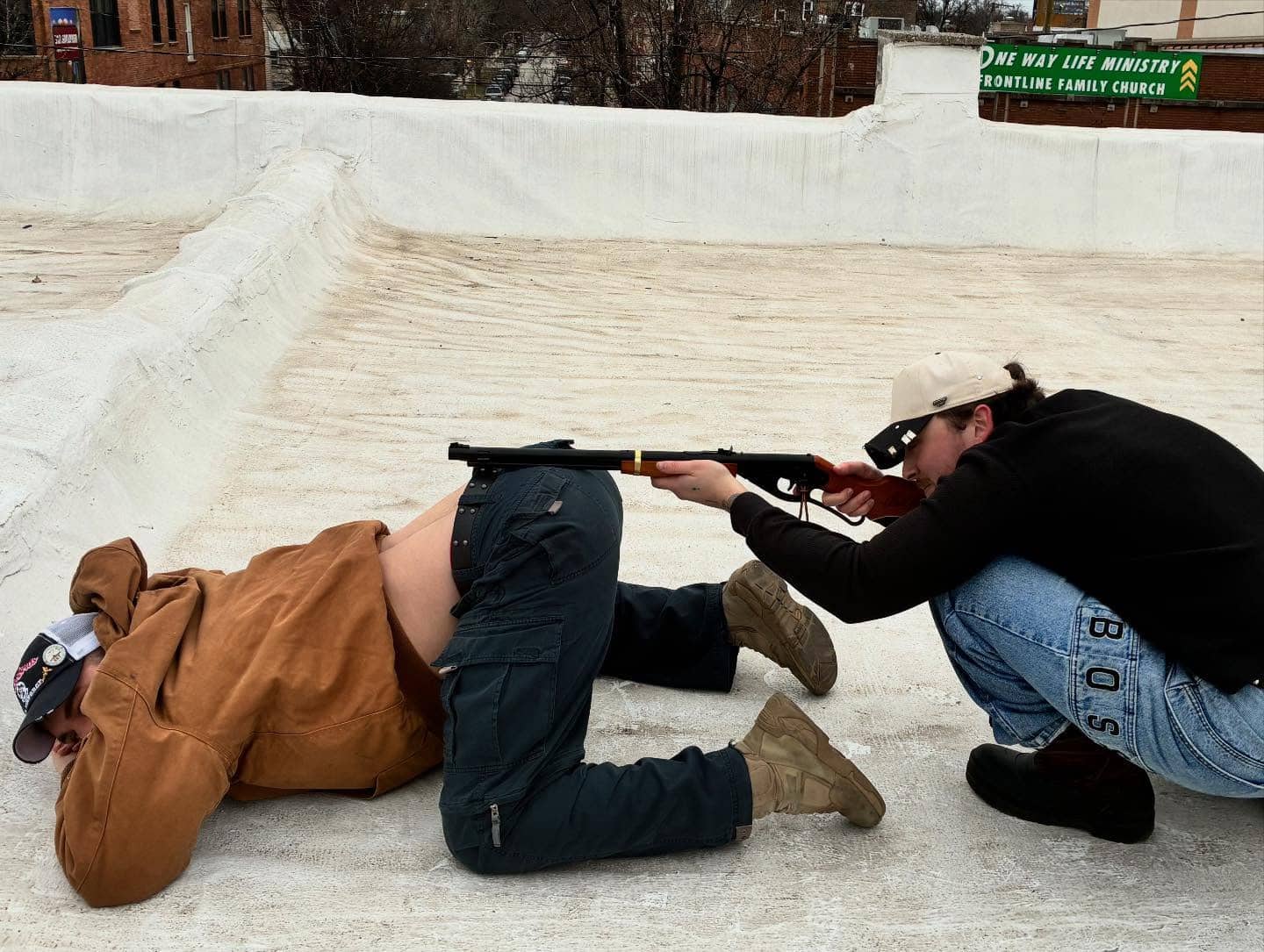 Two men on a rooftop using one person’s back as a literal gun rest.