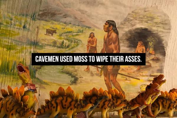 A diorama of early humans near a cave with toy dinosaurs placed in the foreground, including a surprising historical fact that cavemen used moss as a primitive form of toilet paper.