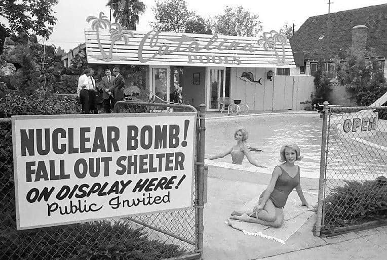 Women in swimsuits lounge by a pool with a sign for a nuclear bomb fallout shelter, a surreal historical photo.
