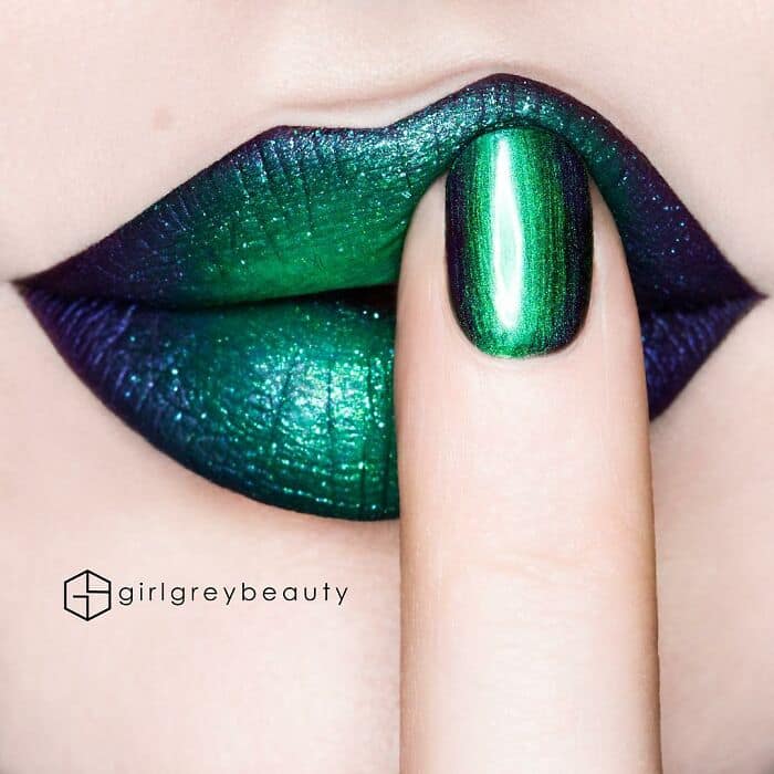 Mesmerizing and amazing lip art featuring a metallic duochrome color shift of emerald green and deep purple, perfectly matched to a glossy fingernail resting on the lower lip in a classic Andrea Reed lip art pose.