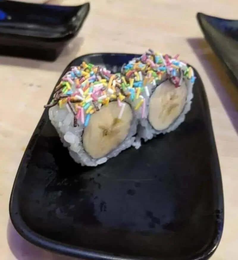 cursed food image from a meme dump showing two pieces of "dessert sushi" where the filling is a raw banana and the topping is a layer of multicolored candy sprinkles on white rice.