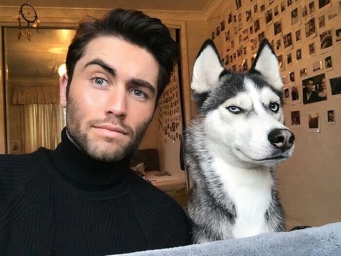 Man in black turtleneck posing next to a husky with matching blue eyes and expression.
