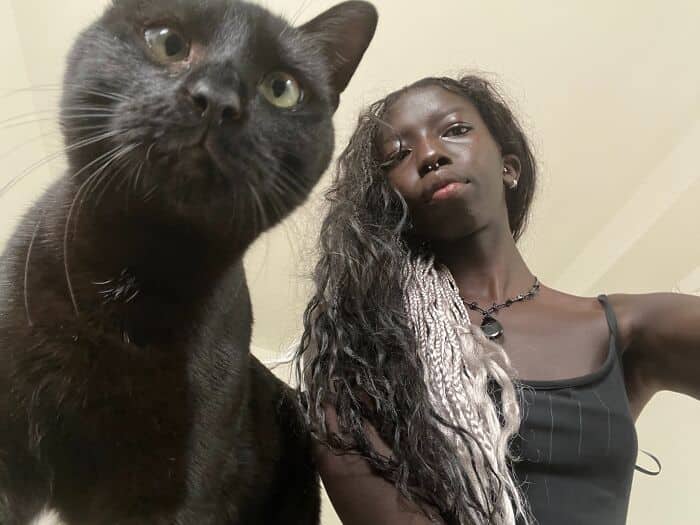 Low-angle selfie of a woman with long dark braids and a sleek black cat looking identical.