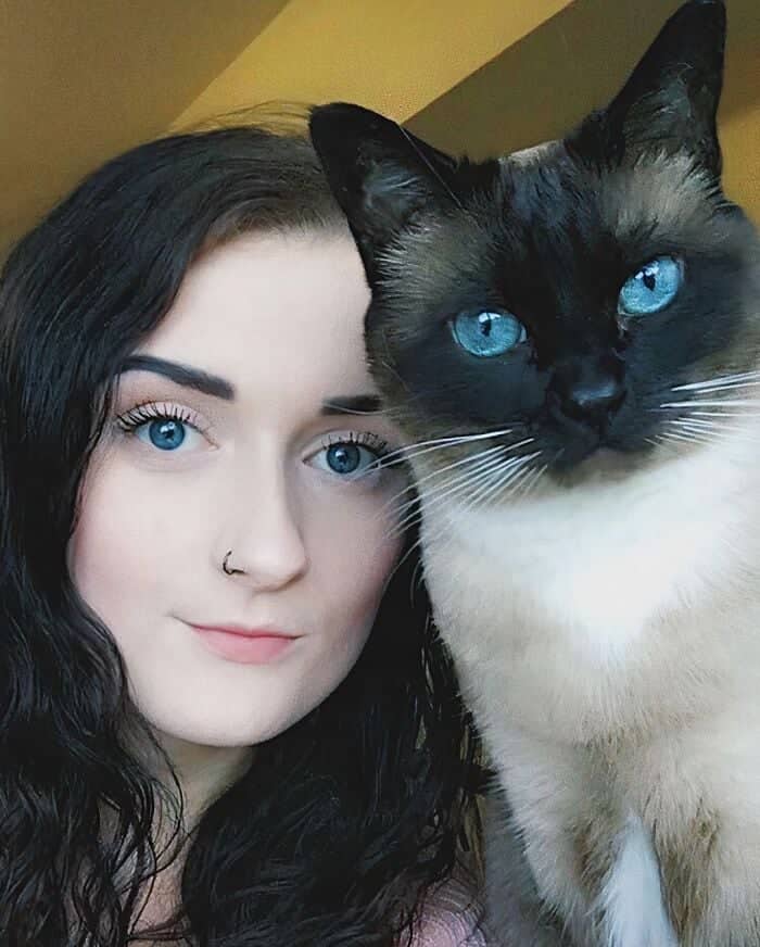 A side-by-side portrait of a woman and her Siamese cat both featuring striking bright blue eyes.
