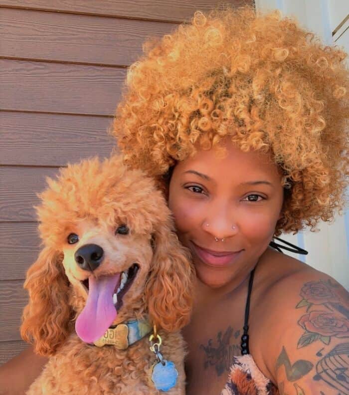 Smiling woman with a blonde afro posing with a toy poodle having similar curly fur.