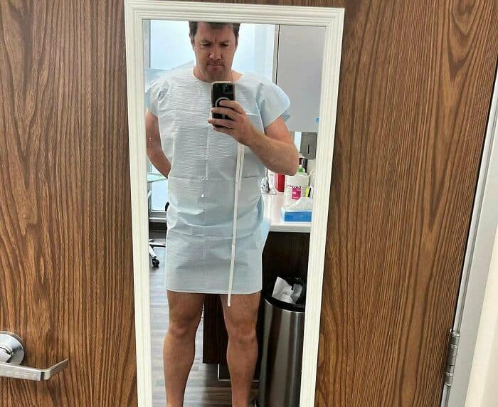 A tall people problems selfie of a man in a hospital gown that looks like a mini skirt on his long legs.