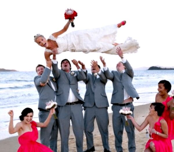 Groomsmen throwing terrified bride into the air on beach for group photo.