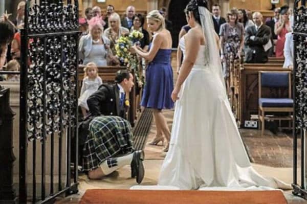 Groom in kilt kneeling to look up under bride's wedding dress in church.