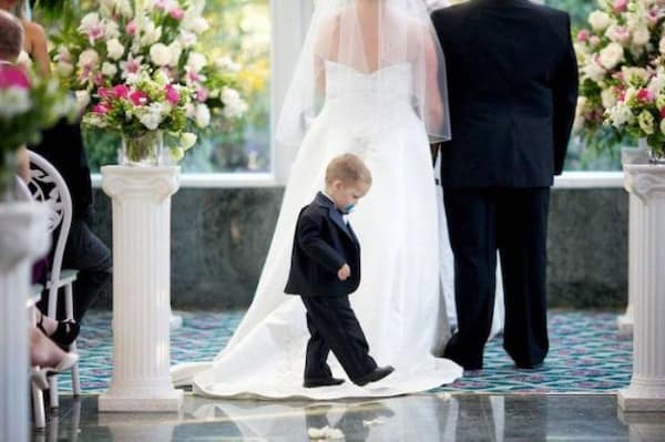 Toddler in tuxedo walking down aisle looking dejected with pacifier in mouth.
