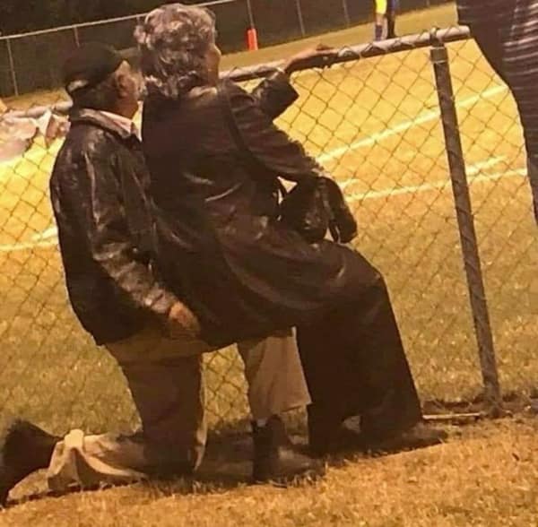 moving wholesome image of an elderly couple at a sports field, where the husband has knelt down to let his wife sit on his knee so she can comfortably watch the game through the chain-link fence.