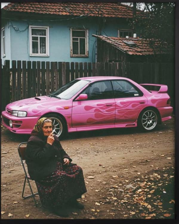 A high-contrast wholesome meme showing an elderly woman sitting in a folding chair next to a bright pink sports car with purple flame decals, proving you’re never too old for a cool ride.