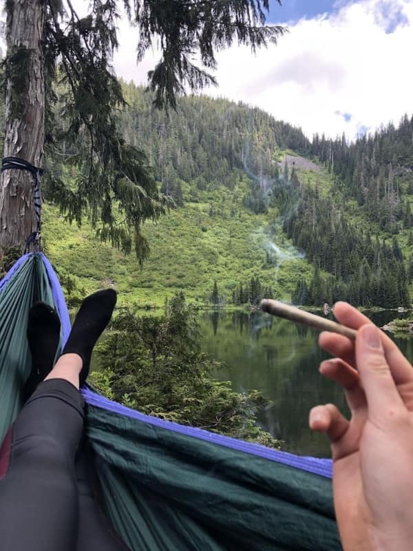 deeply relaxed wholesome image from a first-person perspective in a hammock, overlooking a still mountain lake and forest while holding a joint for a quiet moment of reflection.